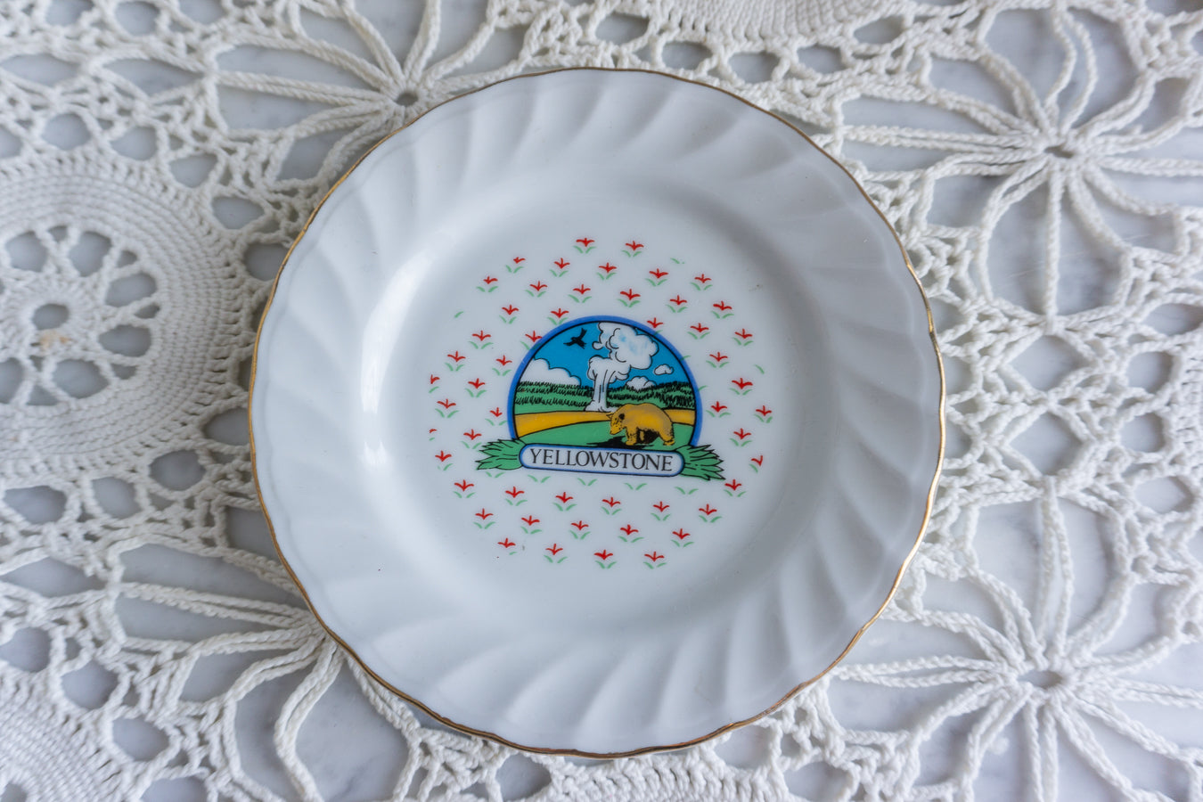 White souvenir plate with Yellowstone National Park illustration, floral background, and gold trim on lace doily