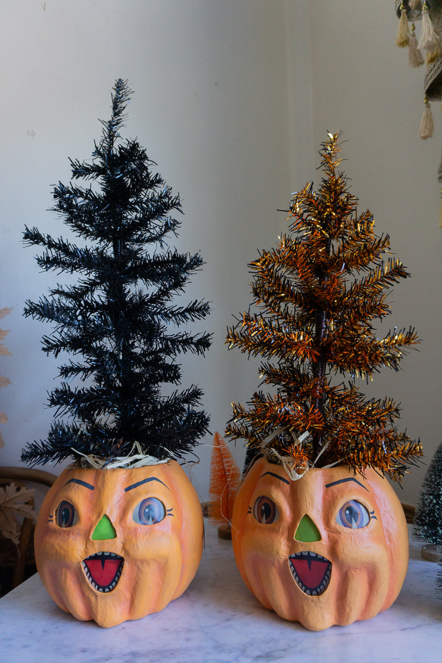 Two pumpkins with face paint and small trees on top, one black and one gold, on a marble surface.